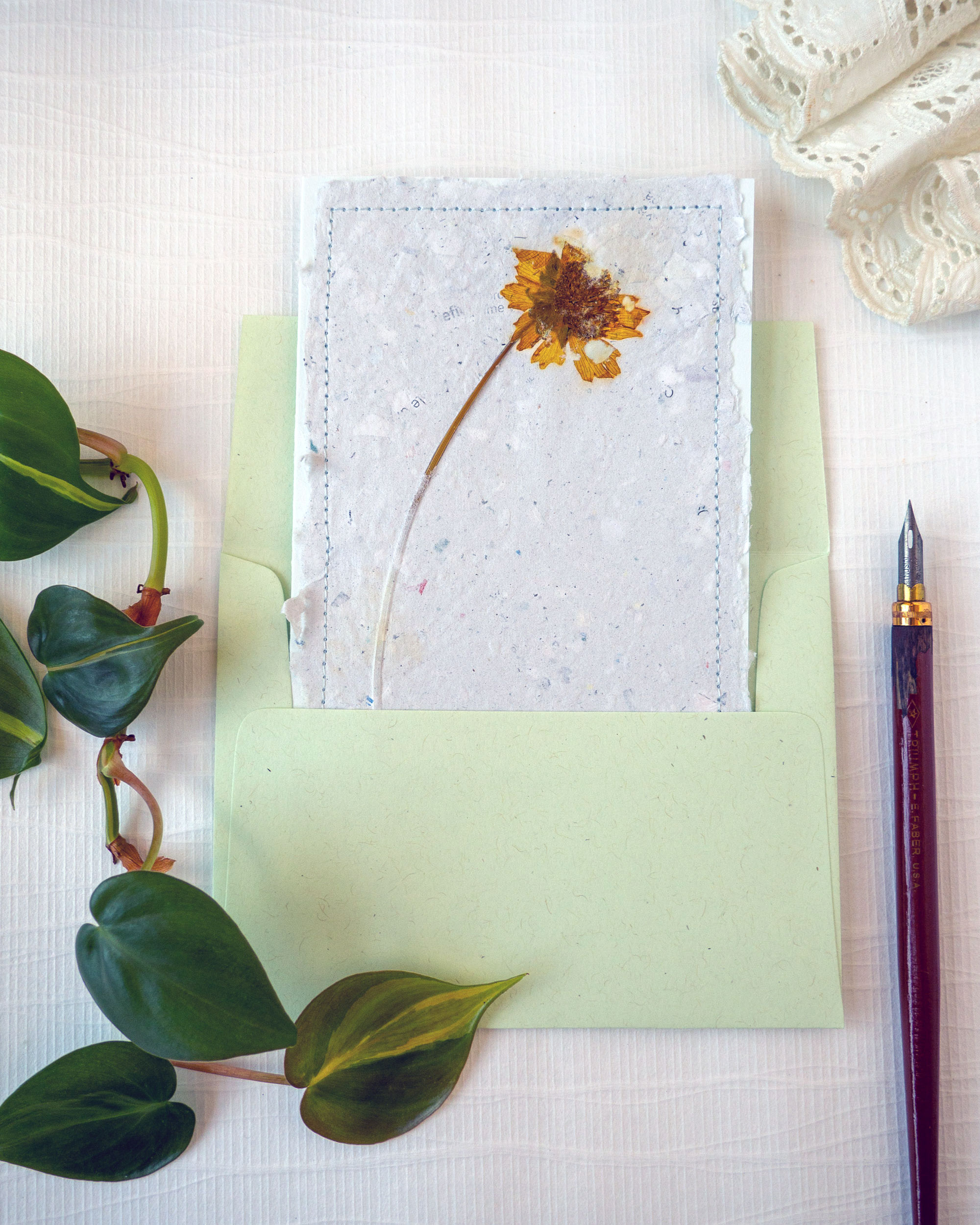 A handmade greeting card featuring handmade paper embedded with a dried yellow flower and stitching around the edge sits inside a light green envelope. Decorative pothos leaves, a calligraphy pen, and lace surround the greeting card.
